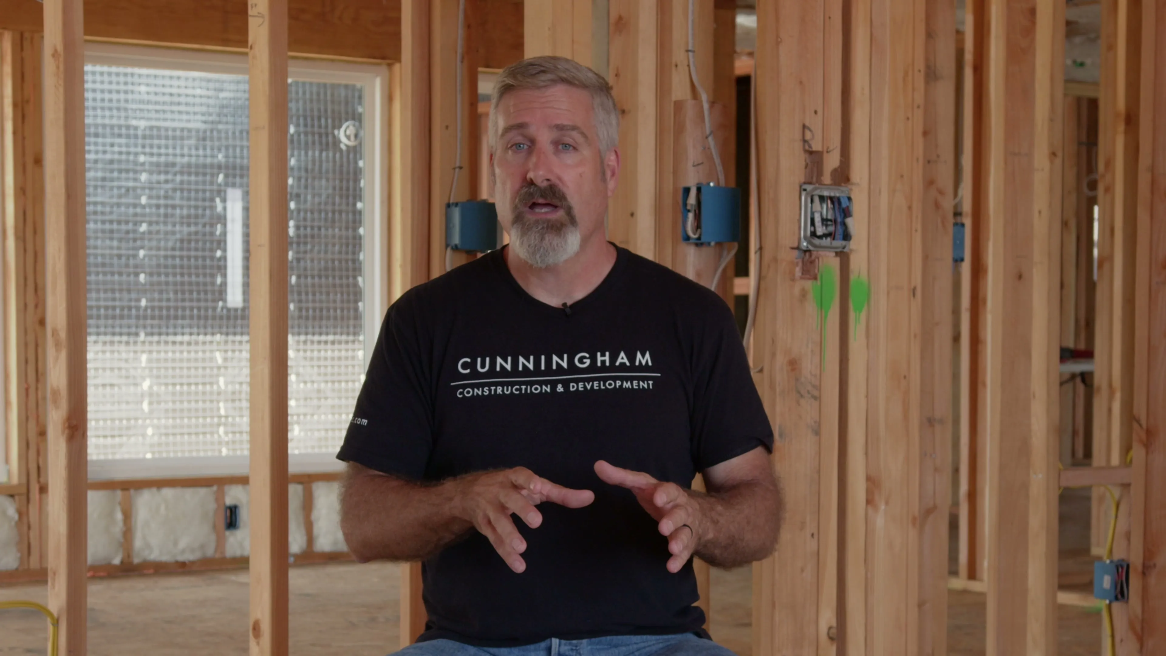 Dustin, in a Cunningham Construction T-shirt, explains with both hands while sitting inside the open wood framing of a new build.